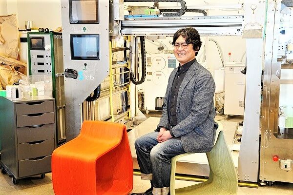 畳二畳分の大きさの大型3Dプリンターとその前に3Dプリンター製の椅子に腰かける田中浩也教授